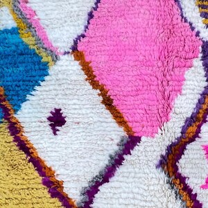 May include: Close-up of a colorful, textured rug with a geometric pattern. The rug features diamond shapes in white, pink, blue, and yellow, outlined with purple and brown. The rug has a plush, soft appearance.