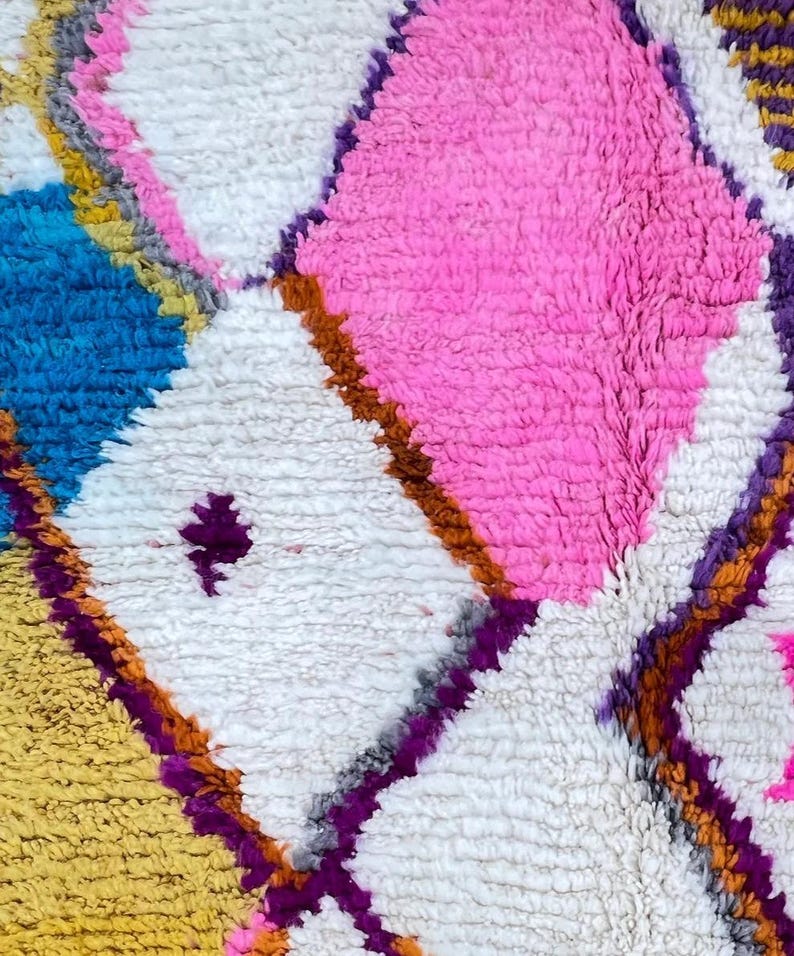 May include: Close-up of a colorful, textured rug with a geometric pattern. The rug features diamond shapes in white, pink, blue, and yellow, outlined with purple and brown. The rug has a plush, soft appearance.