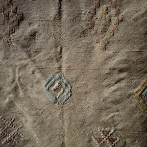 May include: Close-up of a beige textile with embroidered geometric patterns. The design includes diamond and cross shapes in shades of brown, blue, and yellow. The fabric has a textured appearance.