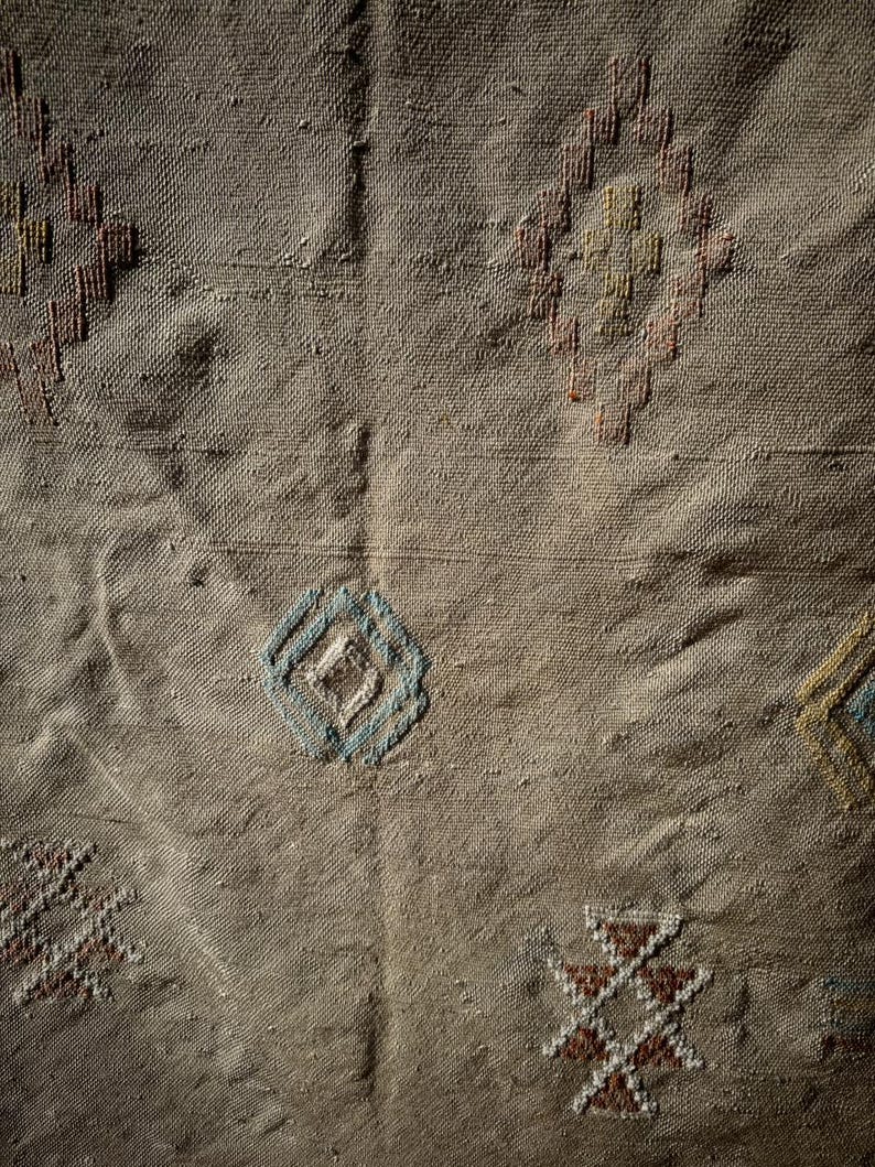 May include: Close-up of a beige textile with embroidered geometric patterns. The design includes diamond and cross shapes in shades of brown, blue, and yellow. The fabric has a textured appearance.
