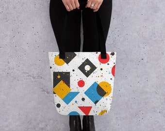 May include: A white tote bag with a geometric pattern featuring black, yellow, red, and blue shapes. The pattern is on a white background with a splatter of white paint.