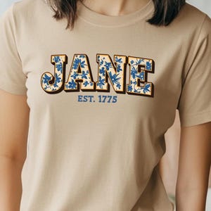May include: A beige t-shirt with a floral design in blue and yellow. The text "JANE EST. 1775" is printed on the front of the shirt.