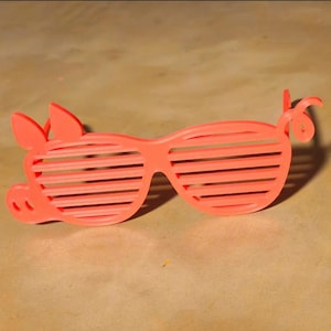 May include: Coral-colored novelty sunglasses shaped like a pig. The sunglasses feature horizontal slats for lenses and pig-like details, including ears, a snout, and a curly tail. The glasses are set against a neutral background.