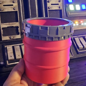 Peut inclure: Un récipient cylindrique rouge avec un bord gris. Le récipient est tenu dans une main, avec un arrière-plan détaillé d'un panneau de commande avec des boutons et des lumières. L'objet semble être imprimé en 3D.