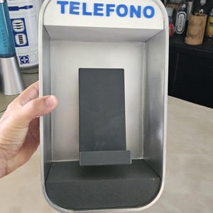 Può includere: Un telefono a muro bianco con la scritta "TELEFONO" in lettere blu sul davanti. Il telefono ha un supporto per telefono nero all'interno.