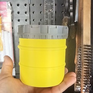 Peut inclure: Un récipient jaune vif avec un couvercle gris est tenu dans une main. Le récipient a une forme cylindrique avec des rainures horizontales. Des outils et une règle de mesure sont visibles en arrière-plan.