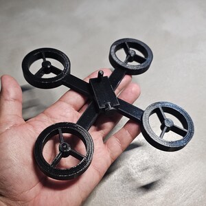 May include: A black plastic drone frame with four propellers. The frame is designed to hold a drone motor and propeller on each arm.