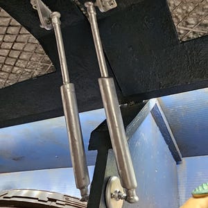 Könnte beinhalten: Zwei silberfarbene Gasdruckfedern sind an einer schwarzen Holzkonstruktion befestigt. Die Streben sind mit silbernen Halterungen und kreisförmigen Halterungen befestigt. Der Hintergrund zeigt ein blaues und schwarzes Interieur.
