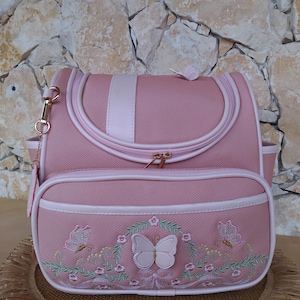 Könnte beinhalten: Rosafarbene Reisetasche mit Schmetterlingsdesign. Die Tasche hat einen oberen Griff, eine vordere Reißverschlusstasche und einen goldfarbenen Reißverschluss. Die Vordertasche hat gestickte Schmetterlinge und florale Akzente. Die Tasche ist mit weißem Zierrat versehen.