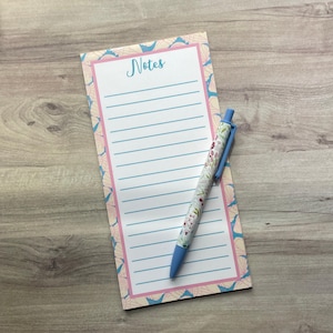 May include: A rectangular notepad with a white center featuring the word Notes in blue script, surrounded by a border of pink and blue seashells. A retractable ballpoint pen with a floral pattern and a blue clip rests diagonally on the notepad.