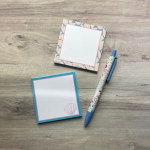 May include: Two square sticky note pads and a retractable ballpoint pen. One pad features a border of seashells, while the other has a solid blue border and a single seashell icon in the corner. The pen has a floral pattern and a blue clip.