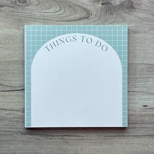 May include: A square notepad with a light blue grid pattern and a white arched section. The words "THINGS TO DO" are printed in a dark blue font across the top of the white section. The notepad is on a wooden surface.