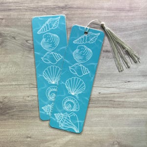 May include: Two rectangular bookmarks with a teal background featuring white line art illustrations of various seashells. The bookmarks have a smooth finish and one includes a silver-colored tassel attached through a hole at the top edge.