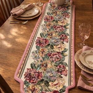 Peut inclure: Chemin de table en tapisserie florale avec un fond crème et un motif répété de fleurs roses, rouges et bleues. Le chemin de table a une bordure rose festonnée et est posé sur une table en bois. La table est dressée avec des assiettes, des serviettes et de la verrerie.