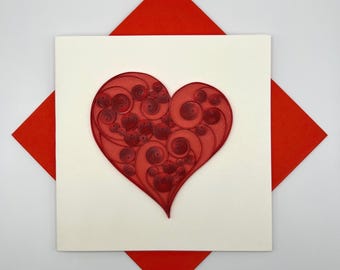 May include: A white card with a red paper quilled heart design. The heart is made of many small, curled paper pieces.