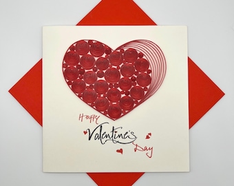May include: A Valentine's Day card featuring a red paper heart design on an off-white background. The heart is crafted from rolled paper. The card rests on a red envelope. The words "Happy Valentine's Day" are inscribed on the card.