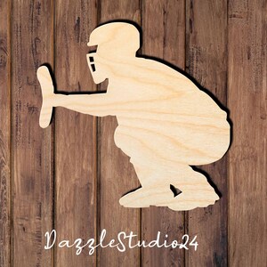 May include: A light-colored wooden cutout silhouette of a baseball catcher in a crouched position, wearing a helmet and face mask, with one arm extended forward. The text DazzleStudio24 is printed in white script at the bottom of the image.