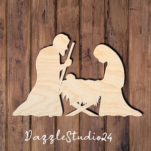 May include: A light-colored wooden cutout depicting a nativity scene with two figures kneeling on either side of a central manger containing a small infant. The figures are rendered in a flat, silhouette style with visible wood grain texture.