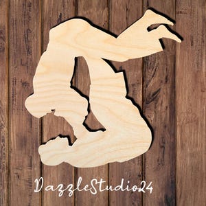 May include: A light-colored wooden cutout featuring the silhouette of a person performing a backflip or gymnastic maneuver. The natural wood grain is visible across the surface of the shape. The text DazzleStudio24 is printed in white at the bottom.
