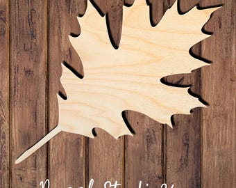 May include: A light-colored, flat wooden cutout shaped like a large oak leaf with distinct pointed lobes and a thin stem. The natural wood grain is visible across the surface of the leaf, which is positioned against a dark brown wood plank background.