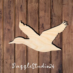 May include: A flat, light-colored wooden cutout in the shape of a duck in flight with wings extended. The surface shows a natural wood grain pattern. The text DazzleStudio24 is printed in white script at the bottom of the wooden duck shape.
