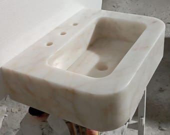 May include: White marble bathroom sink with a rectangular shape and rounded edges. The sink has three holes on the top for faucets and a single drain hole in the bottom.