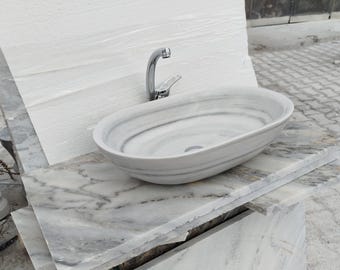 May include: Oval-shaped marble sink with a chrome faucet. The sink is white with gray veining and sits on a stack of marble slabs. The faucet is curved and modern, adding a sleek touch to the bathroom fixture.