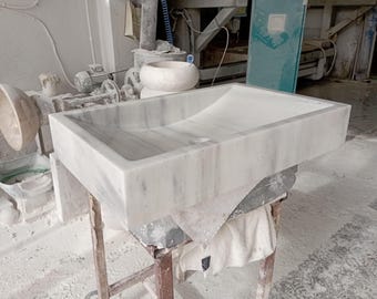 May include: A rectangular marble sink with a gently curved interior. The sink is white with gray veining, and sits on a wooden stand. The sink is a bathroom fixture.