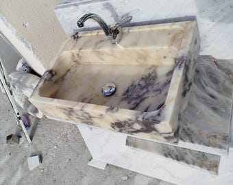 May include: A white marble bathroom sink with a silver tap and a drain stopper. The sink is rectangular and wall-mounted.