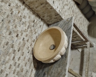 May include: A beige stone sink with a decorative edge sits on a countertop made of beige stone tiles. The sink has a drain in the center.