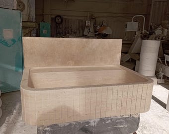 May include: A large, rectangular, stone sink with a backsplash. The sink is made of a light beige stone with a textured surface. The sink is sitting on a black surface.