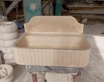 May include: A large, rectangular, stone sink with a decorative, fluted edge. The sink is made of a light beige stone with a smooth, polished finish. The sink has a backsplash with a curved top edge.