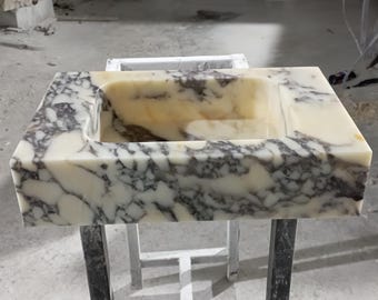 Könnte beinhalten: Ein weißes Marmorwaschbecken mit grauen Adern. Das Waschbecken ist rechteckig und hat ein eingebautes Becken. Das Waschbecken ist auf einem Metallgestell montiert.