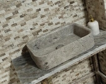 May include: A gray stone rectangular sink with a smooth, slightly textured surface. The sink is set on a countertop with a gray and white mosaic tile backsplash.