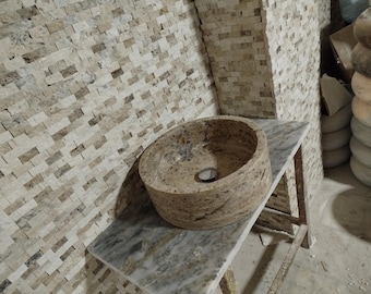 May include: A round stone sink with a rough, textured surface. The sink is set on a white countertop with a brown and white tiled wall behind it.