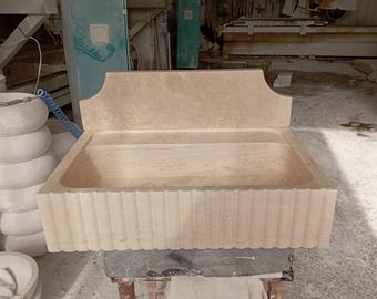 May include: A large, rectangular stone sink with a decorative, fluted edge. The sink is made of light beige marble and has a backsplash with a curved top.
