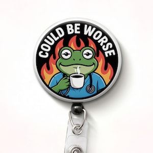 May include: A round badge reel with a cartoon frog wearing a blue medical uniform, holding a coffee cup, and surrounded by flames. The text "COULD BE WORSE" is printed around the edge of the badge. The reel has a black clip and a clear plastic strap.