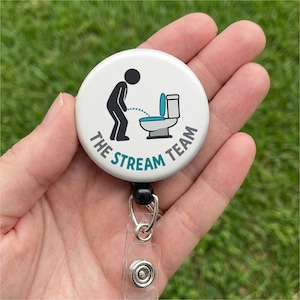 May include: A white badge reel with a black and teal graphic of a person urinating into a toilet. The text "THE STREAM TEAM" is printed around the graphic in teal and gray.