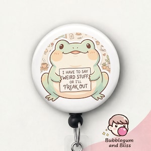 May include: A white badge reel with a cartoon frog holding a sign that reads "I HAVE TO SAY WEIRD STUFF OR I'LL FREAK OUT." The frog is light green with peach accents. The reel has a black retractable cord and a clear plastic strap.