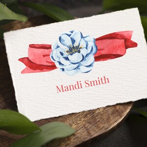 May include: A rectangular white card with deckled edges featuring a watercolor illustration of a light blue flower centered over a horizontal red ribbon. The name Mandi Smith is printed in red serif font below the floral and ribbon graphic.