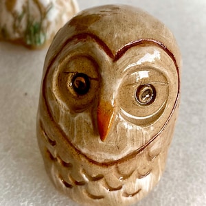 May include: A small, glazed ceramic owl figurine in shades of tan and brown. The owl features a smooth, rounded head with deeply incised circular eyes, a pointed orange beak, and carved feather patterns along the lower body and chest area.