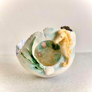 May include: A ceramic bowl featuring a seahorse. The bowl is painted in light blue and white with a textured finish. A yellow seahorse is attached to the side. The interior has green and pink details, and a small seashell.