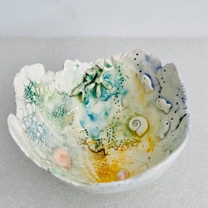 May include: A white ceramic bowl with an irregular, organic rim featuring an interior design of blue, green, and yellow glazes. The bowl is decorated with raised relief elements including small fish, a spiral shell, and coral-like shapes.