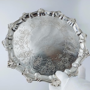 May include: A silver-colored, ornate tray with a scalloped edge and intricate floral patterns. The tray is round and held by a gloved hand. The decorative tray is likely made of metal and features a reflective surface.