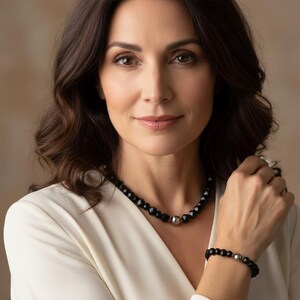 Op de afbeelding: Een vrouw draagt een zwarte kralenketting en armband set. De sieraden hebben facetgeslepen zwarte kralen en zilverkleurige metalen accenten. De vrouw draagt een witte top.