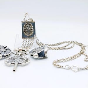 Può includere: Una collana d'argento con più catene e ciondoli. I ciondoli includono un ciondolo quadrato con un design elaborato, un ciondolo a forma di chiave e due ciondoli rotondi con dettagli intricati.