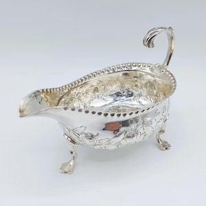 Puede incluir: Una salsera plateada con un asa curva y un pico. El recipiente está decorado con motivos florales y descansa sobre tres pequeños pies. El borde está detallado con pequeños puntos espaciados.