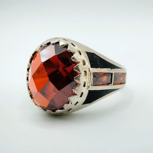 Op de afbeelding: Zilveren ring met een grote, facetgeslepen, ovaal geslepen, rode edelsteen als middelpunt. De ring heeft een zwarte emaille band met drie kleinere rechthoekige rode stenen. De ring heeft een decoratieve, geschulpte rand.