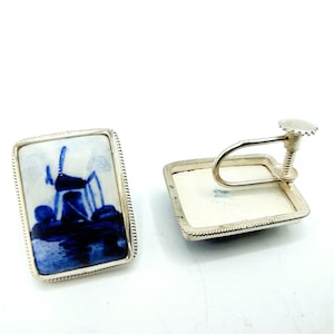 May include: Silver-coloured, rectangular clip-on earrings. Each earring has a blue and white ceramic tile depicting a windmill scene. The tiles are framed with a twisted rope design. The earrings are set against a white background.