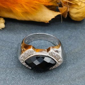 Op de afbeelding: Een zilveren ring met een zwarte edelsteen in een facetgeslepen ontwerp. De ring is versierd met kleine heldere stenen rond de edelsteen.
